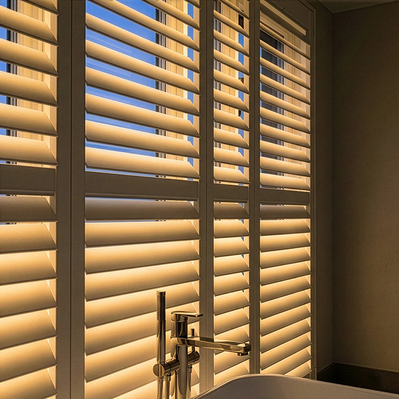 A spa-like bathroom with freestanding tub, softly lit by LiteLüvr shutters at dusk — warm amber glow from the window